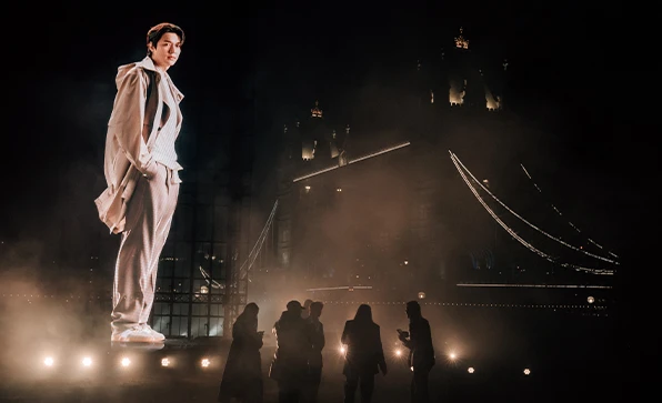 Oversized Lee Minho as a Hologram in front of Tower Bridge in London; there is a group of people on the bottom of the picture (photo)